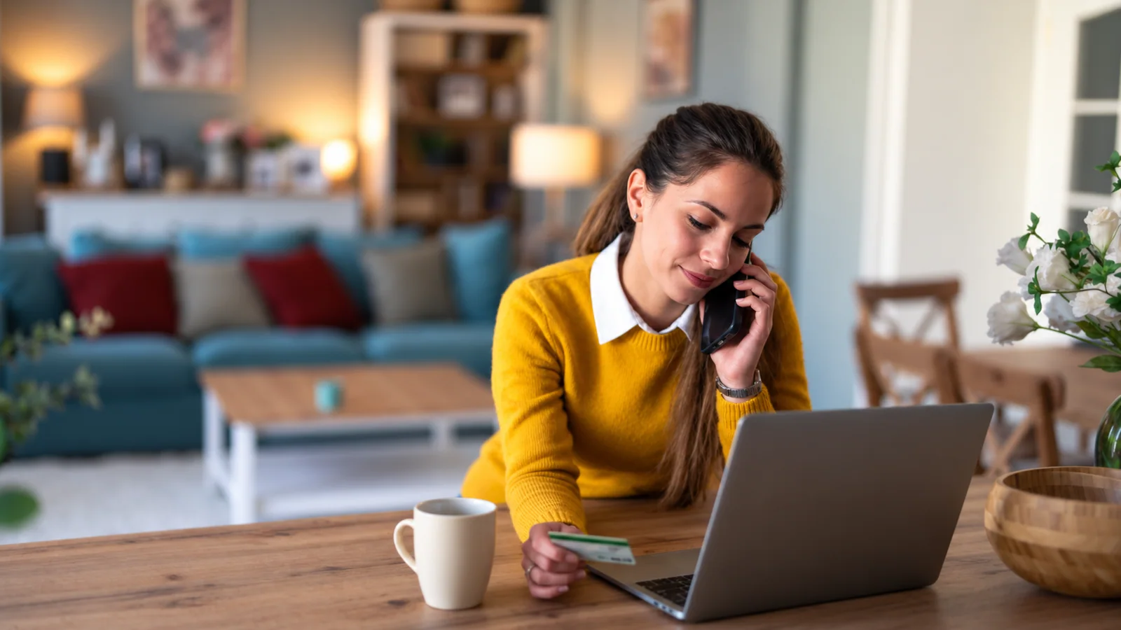 A woman speaking on the phone beside her laptop while holding a card.