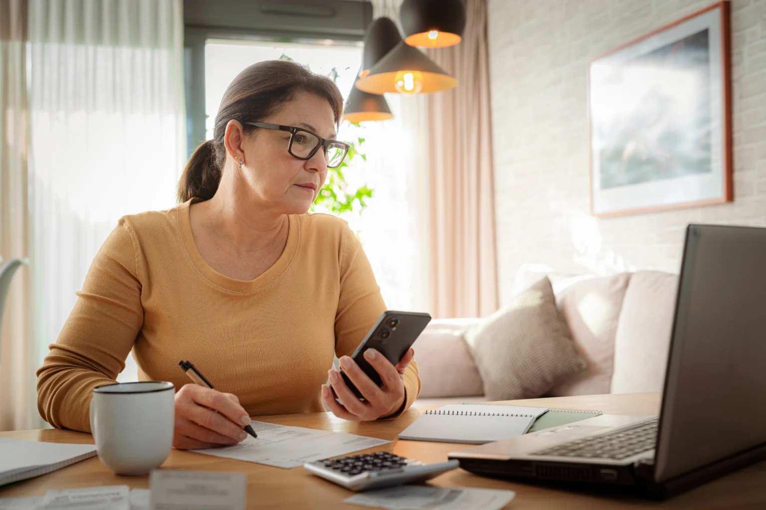 A woman comparing documents with information on her phone and laptop.