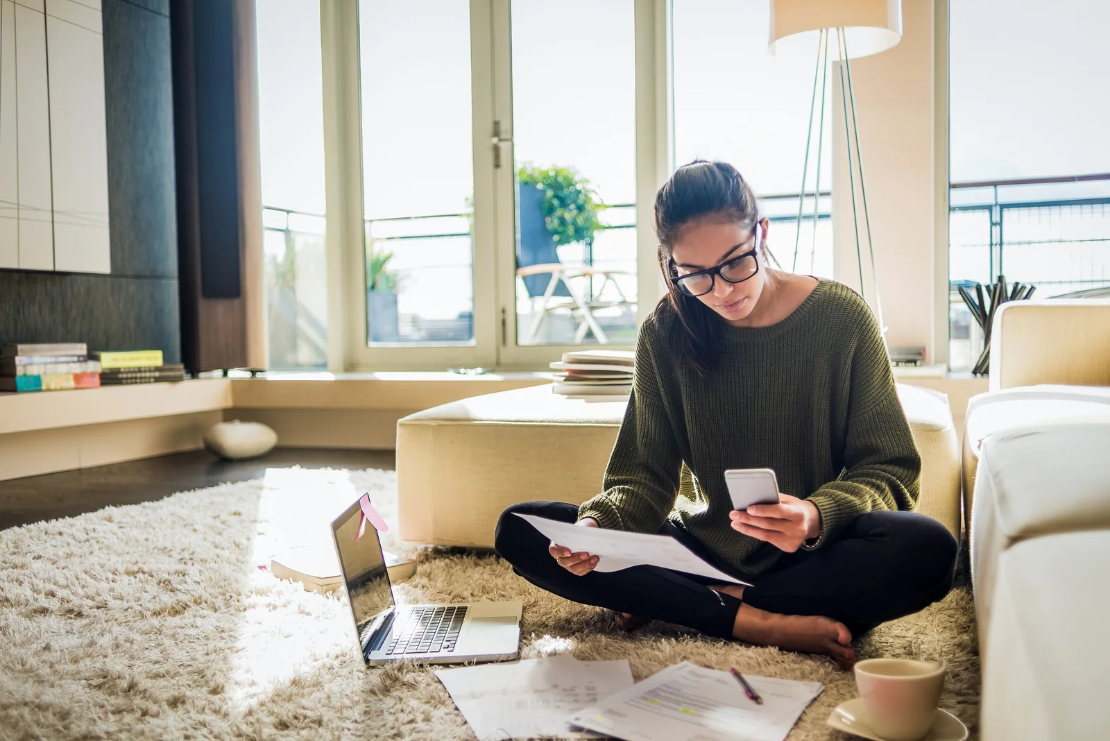 A woman comparing paperwork and her phone beside a laptop at home.
